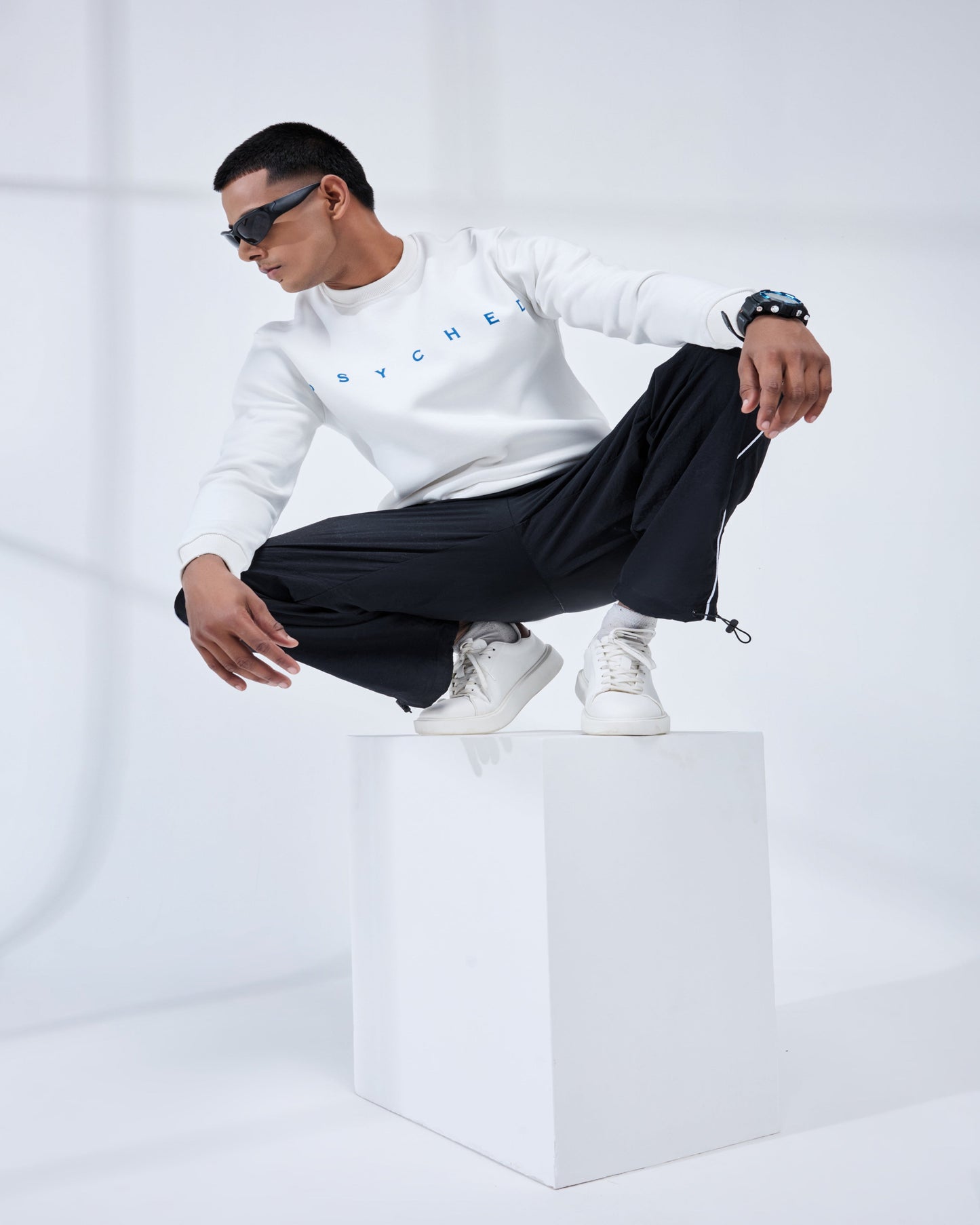 Person wearing a white sweatshirt and black pants sitting on a white cube against a white background