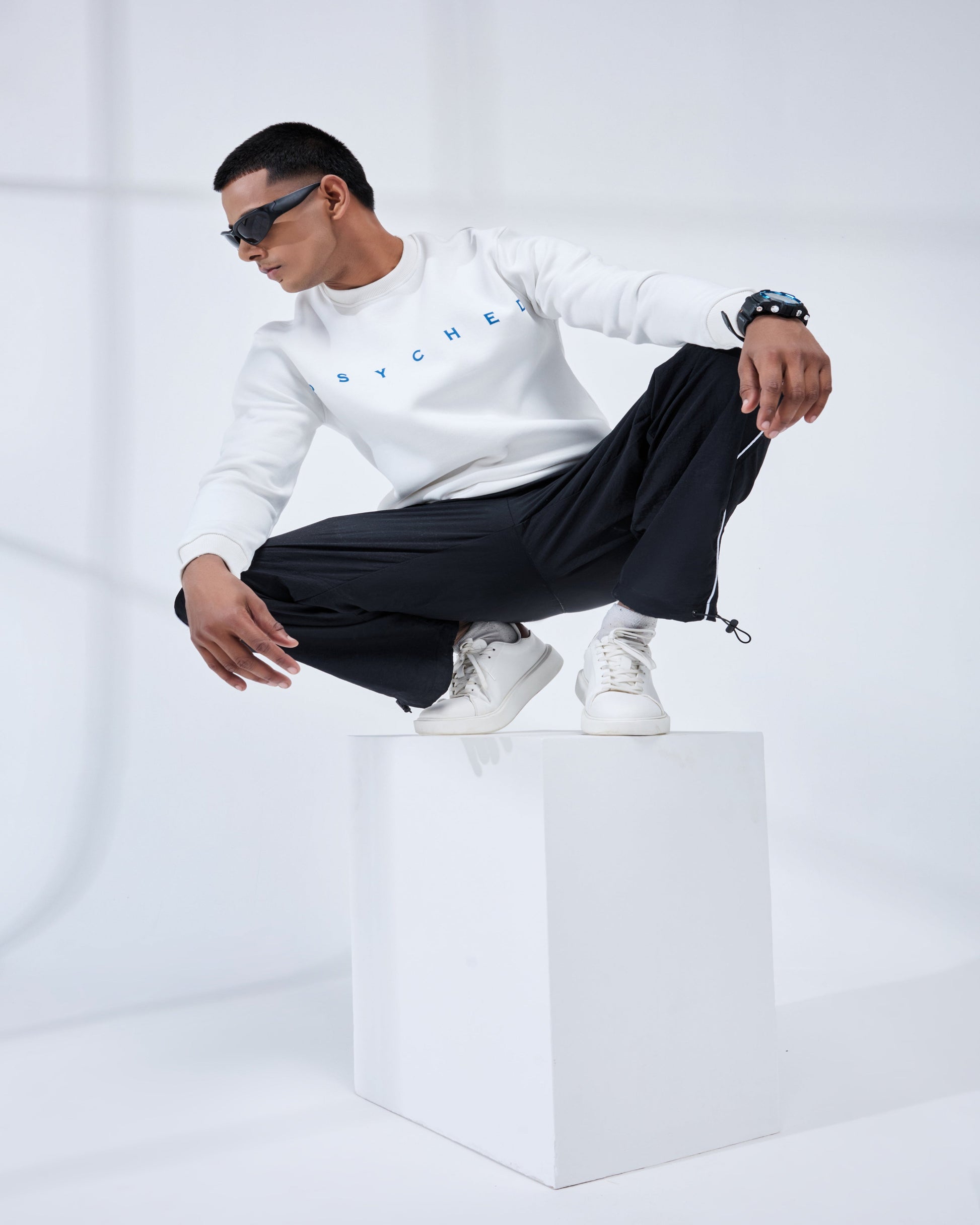 Person wearing a white sweatshirt and black pants sitting on a white cube against a white background