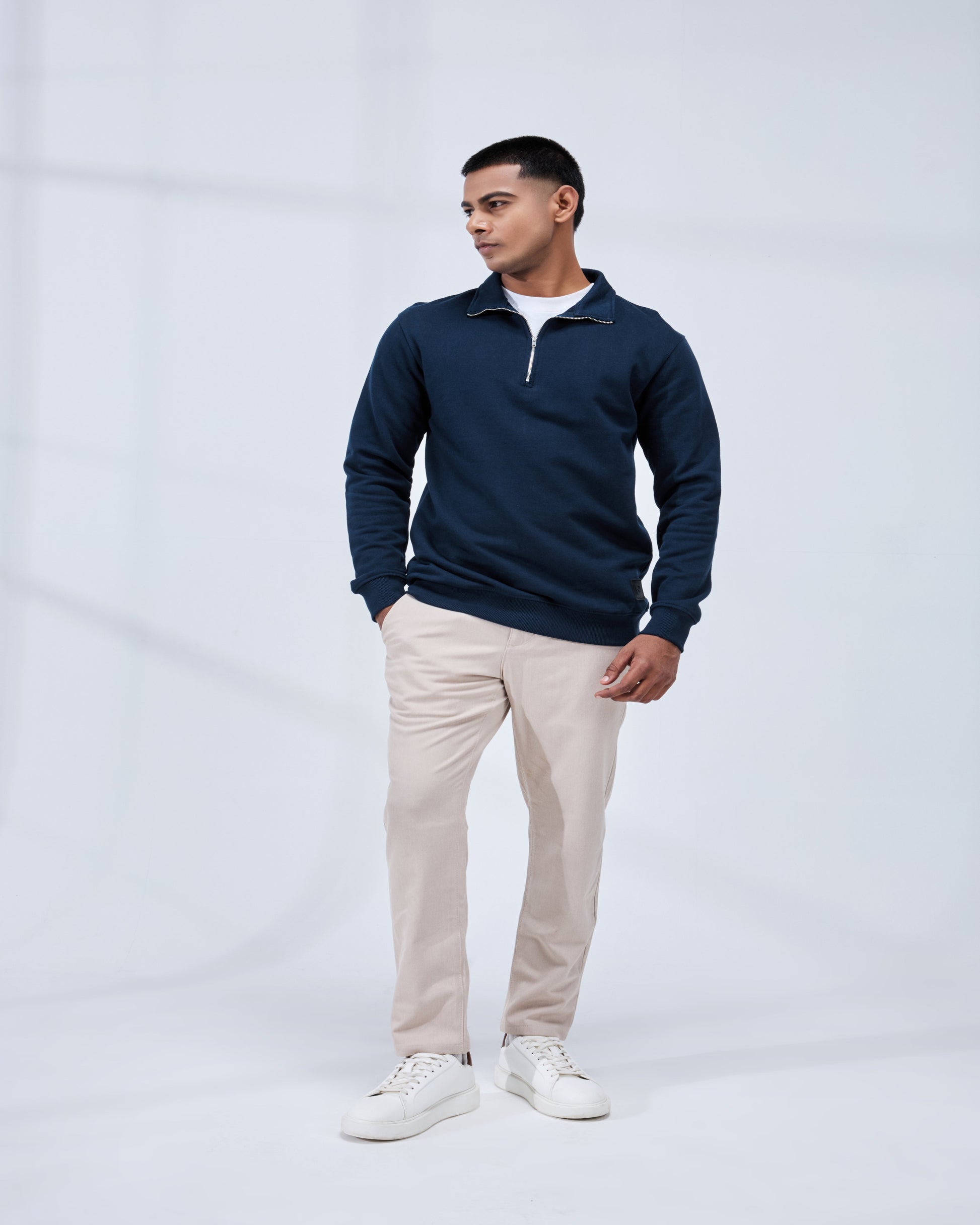 Man wearing a navy blue pullover with a white collar, beige pants, and white sneakers on a light gray background