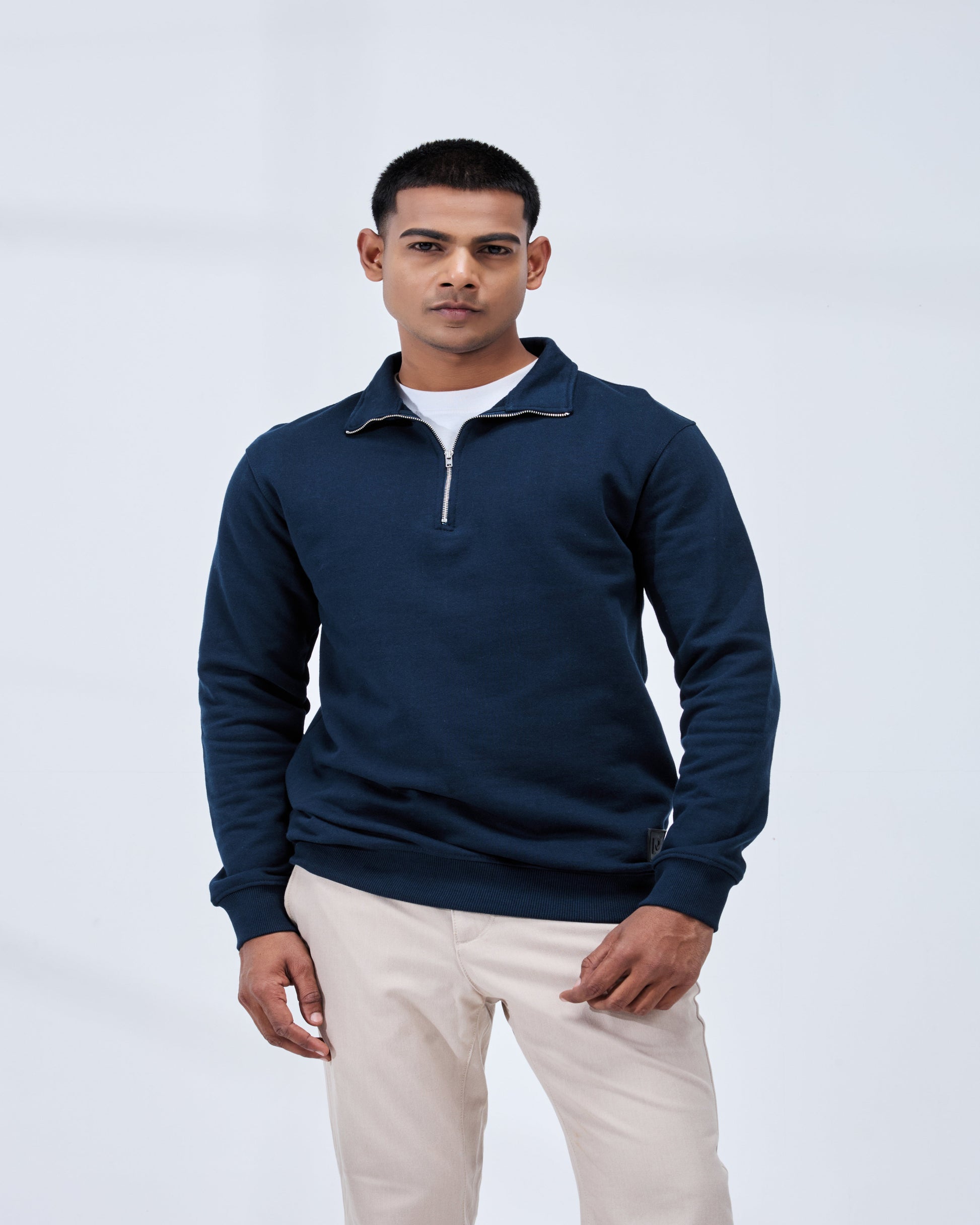 Man wearing a navy blue sweater over a white shirt with beige pants on a light gray background