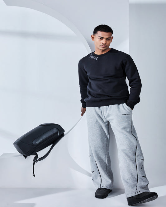 Man in black sweatshirt and gray pants holding a black suitcase against a white background