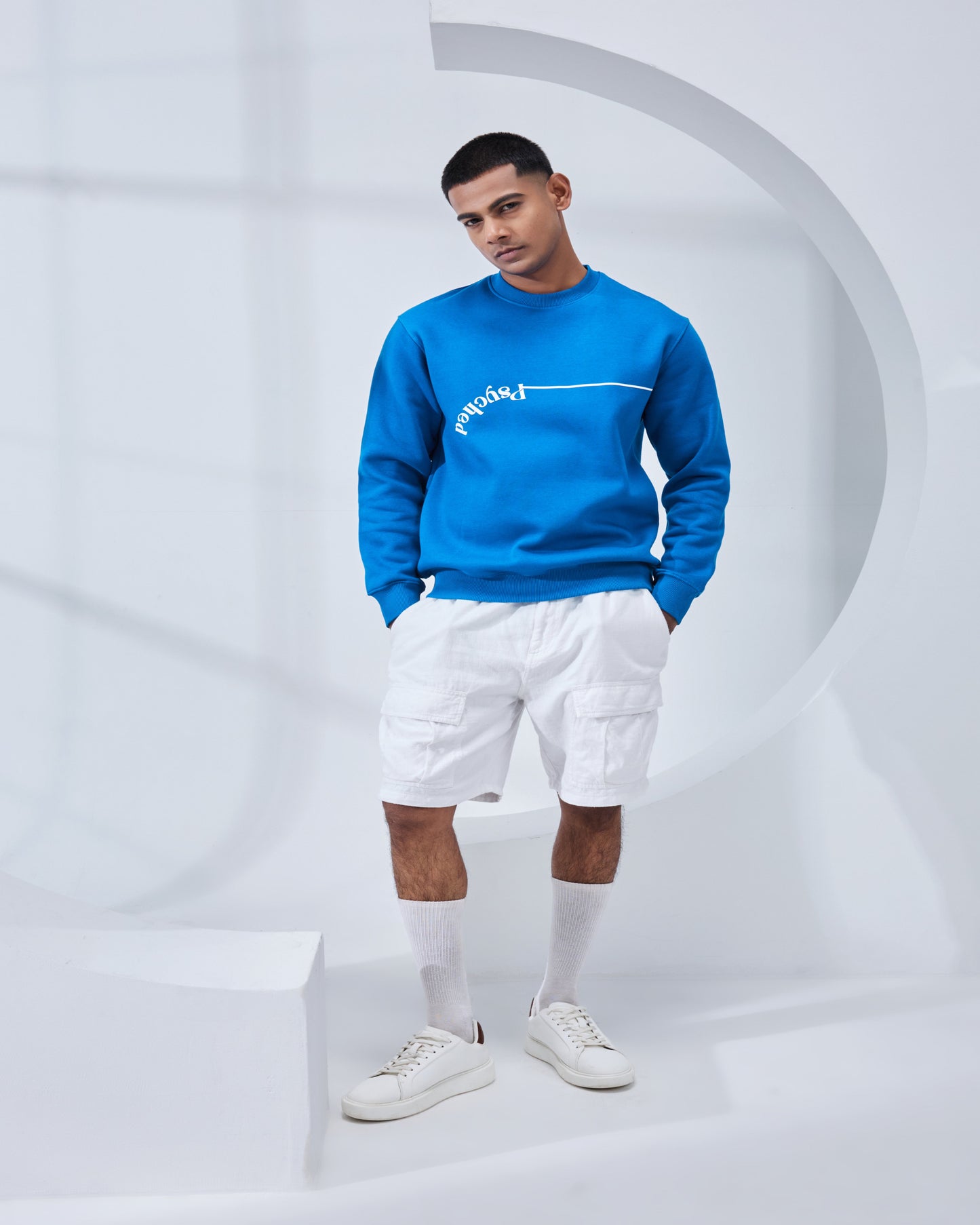 Man wearing a blue sweater, white shorts, and white sneakers against a gray background